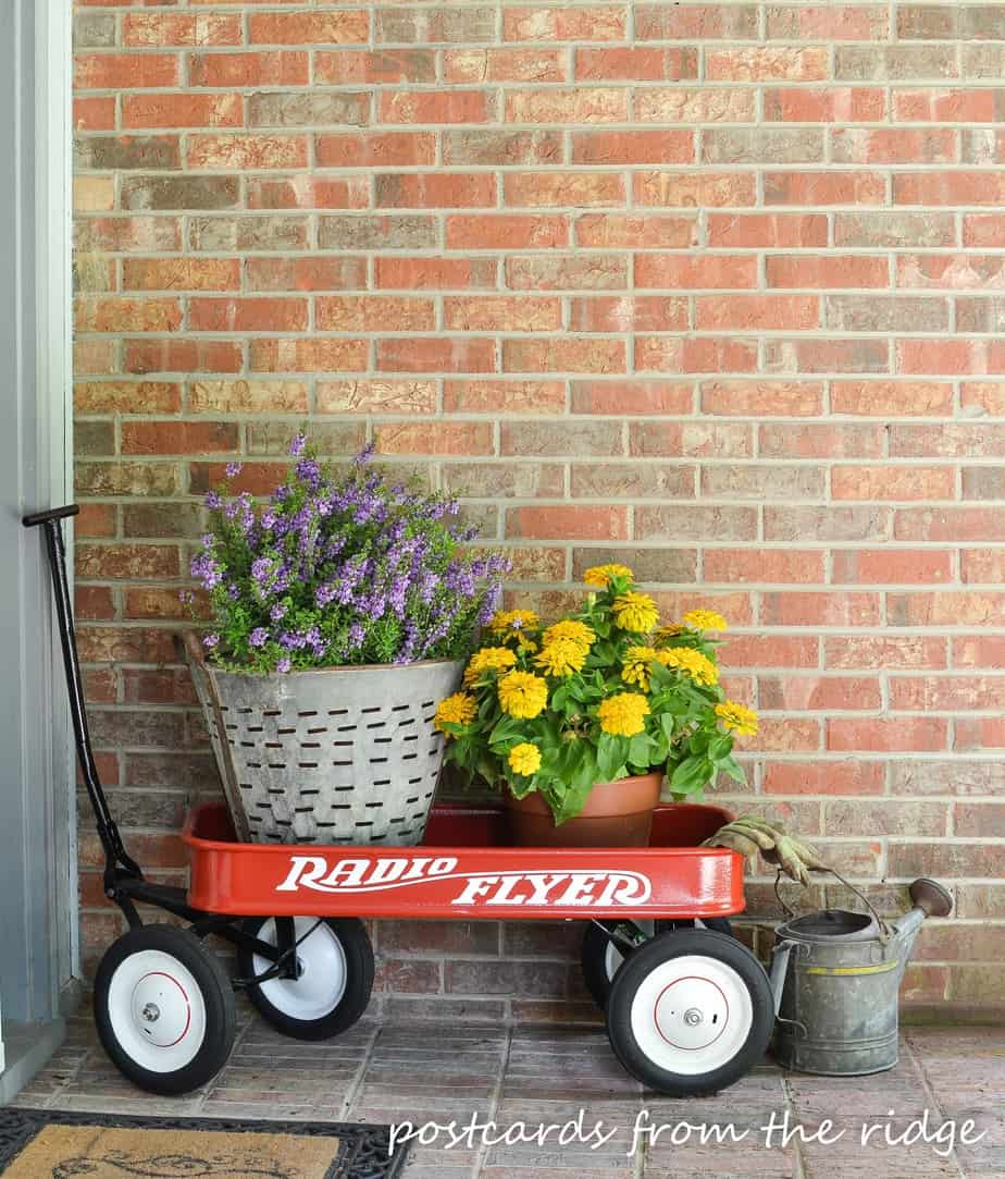 Front porch decor. Vintage Olive Basket with Flowers in a Red Wagon makes an adorable greeting for guests. Postcards from the Ridge.