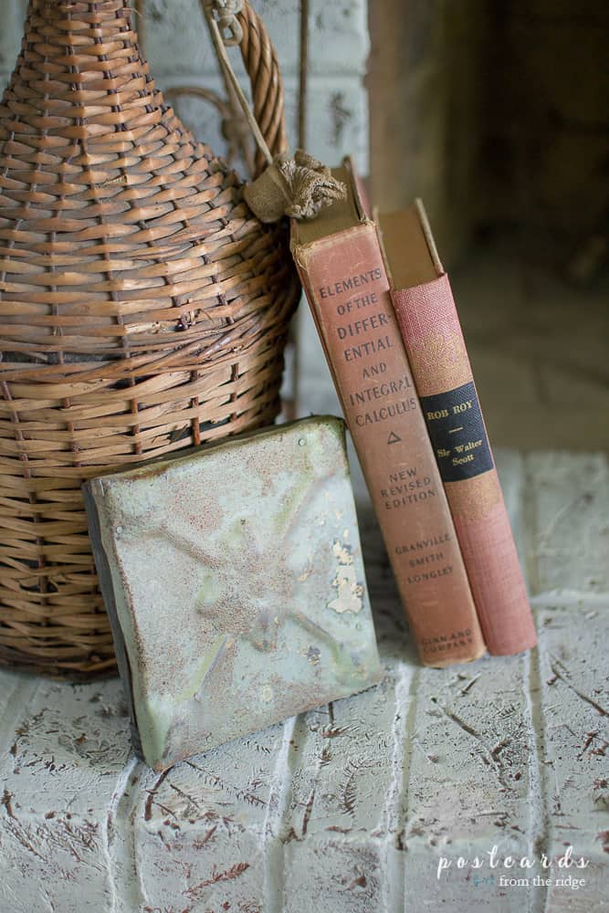 vintage books, ceiling tile plaque, and wicker demijohn on painted brick fireplace hearth