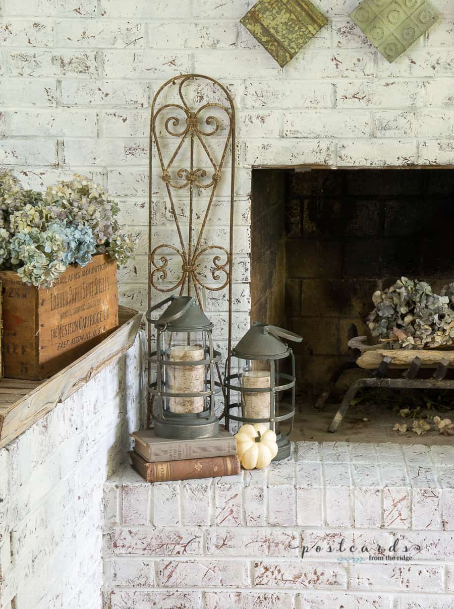 white brick fireplace with dried hydrangeas, old rusty fence section, books, metal lantern, and faux pumpkins