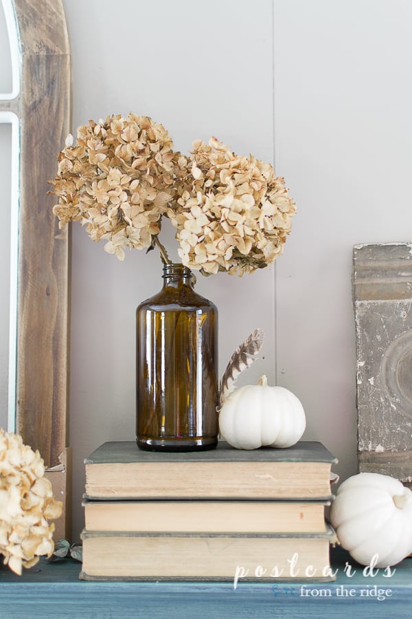 brown bottle with dried hydrangeas and small white pumpkin on fireplace mantel